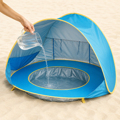A blue beach sun shade tent with yellow trim is set on sand, with a hand pouring water from a clear pitcher into the pool area inside the tent.