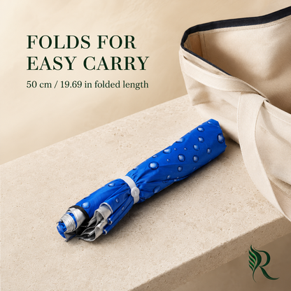 A folded blue umbrella with water droplets on it, placed on a beige stone surface next to a cream canvas tote bag. Text on the image reads 'FOLDS FOR EASY CARRY' and '50 cm / 19.