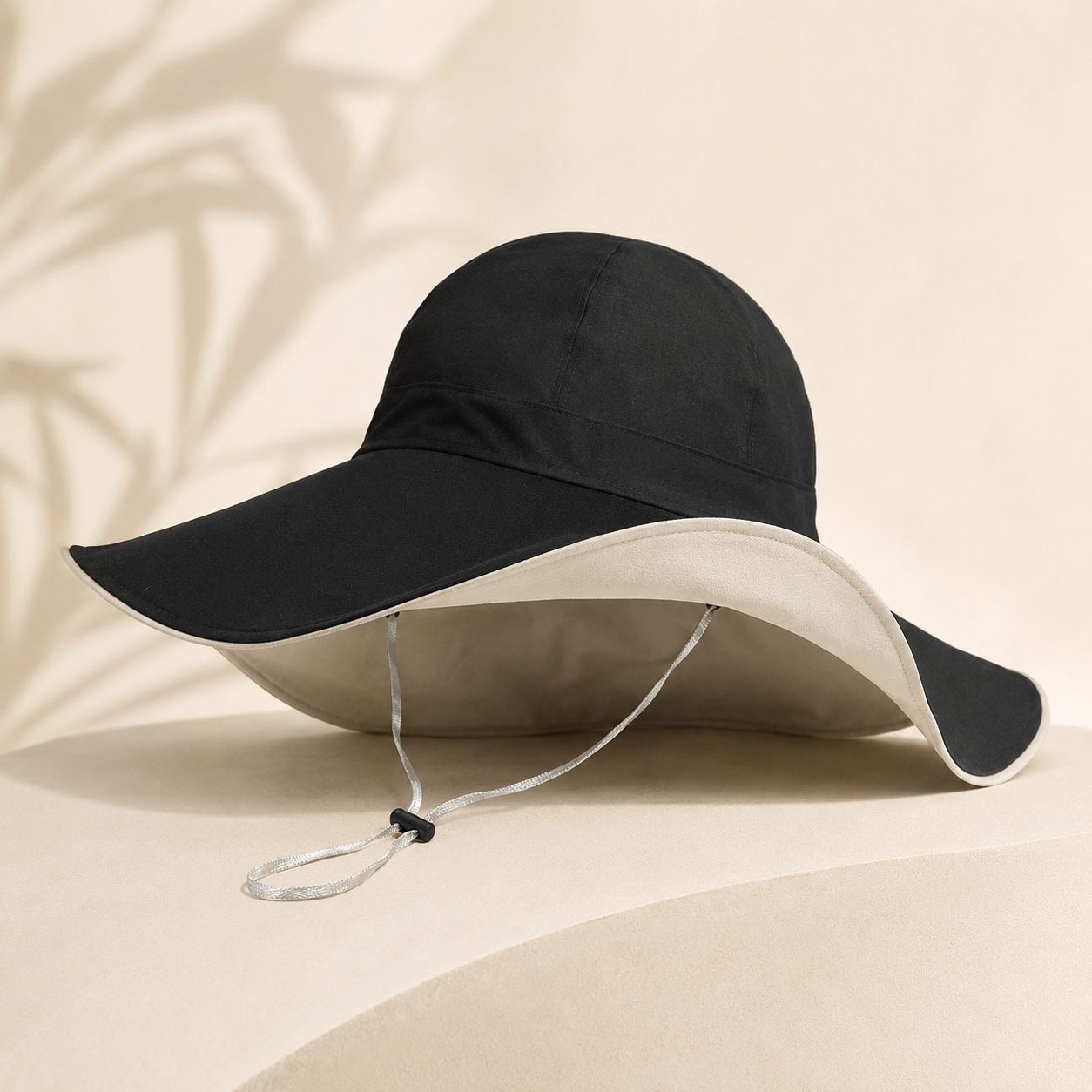 A wide-brimmed sun hat with a black exterior and a light beige interior, featuring a thin chin cord, displayed on a beige surface with soft leaf shadows.