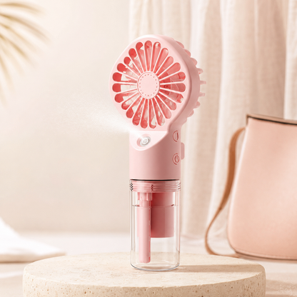 A pink portable handheld fan with a clear water reservoir, shown spraying mist on a beige pedestal with a soft pink bag in the background.