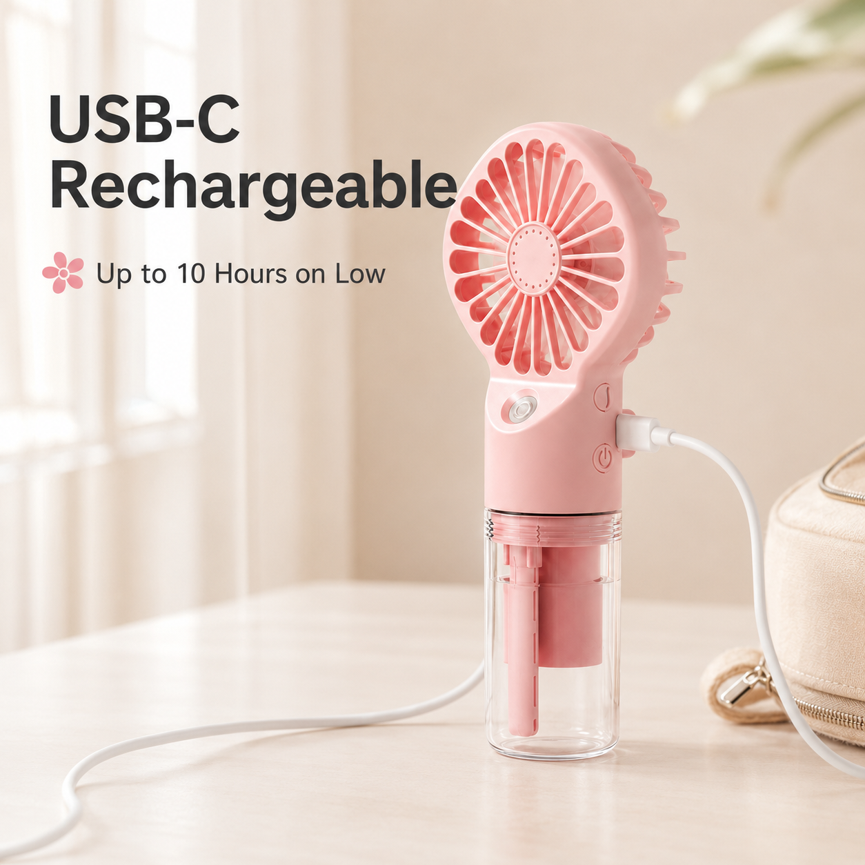 A pink USB-C portable fan with a clear water reservoir sits on a light wooden table, plugged into a white cable. The background is a softly blurred indoor setting with warm beige tones, and text on the left reads 'USB-C Rechargeable' and 'Up to 10 Hours on Low'.