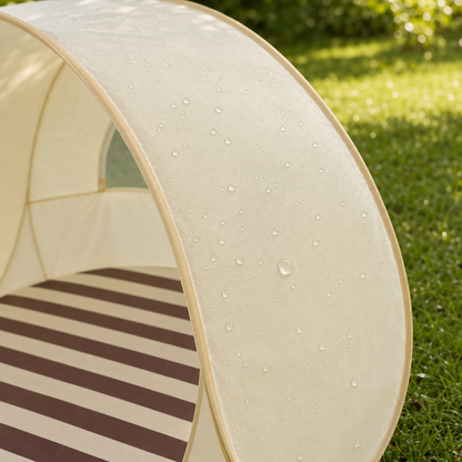 A close-up outdoor shot of a small pop-up canopy tent with a cream-colored fabric roof and a brown-and-cream striped floor, shown on green grass with water droplets on the surface.
