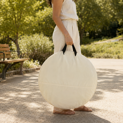 A woman in a white dress carries a large round cream-colored beach umbrella in a sunny park setting.