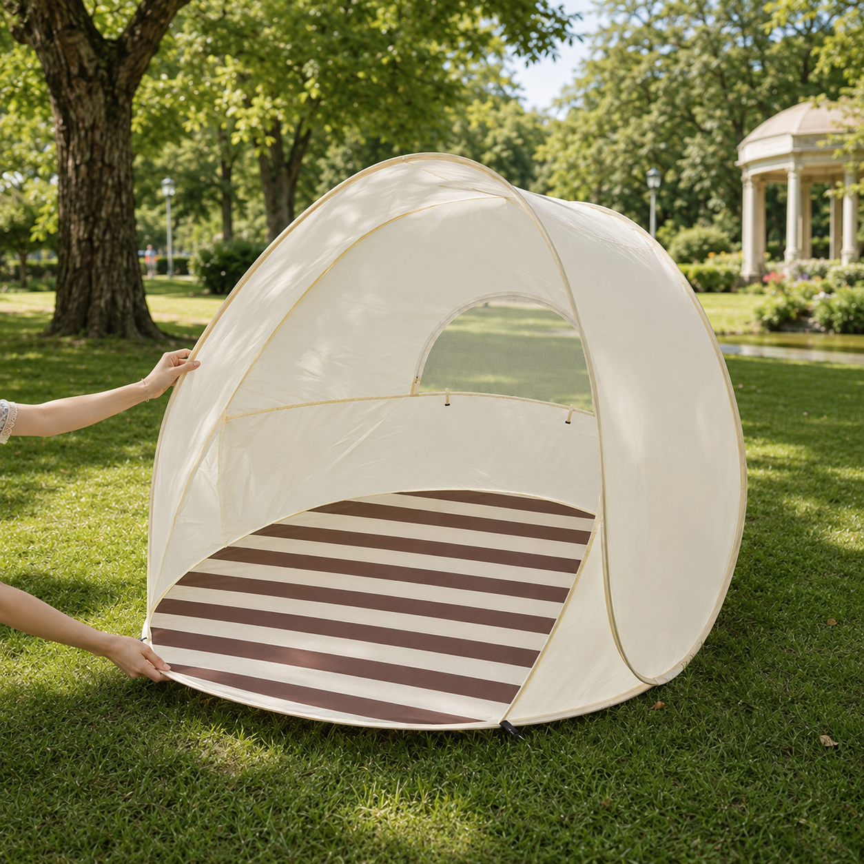 A dome-shaped pop-up tent with a striped floor mat is being set up on a grassy park lawn. A person's hands are visible on the left side holding the tent open, with trees and a gazebo in the background.