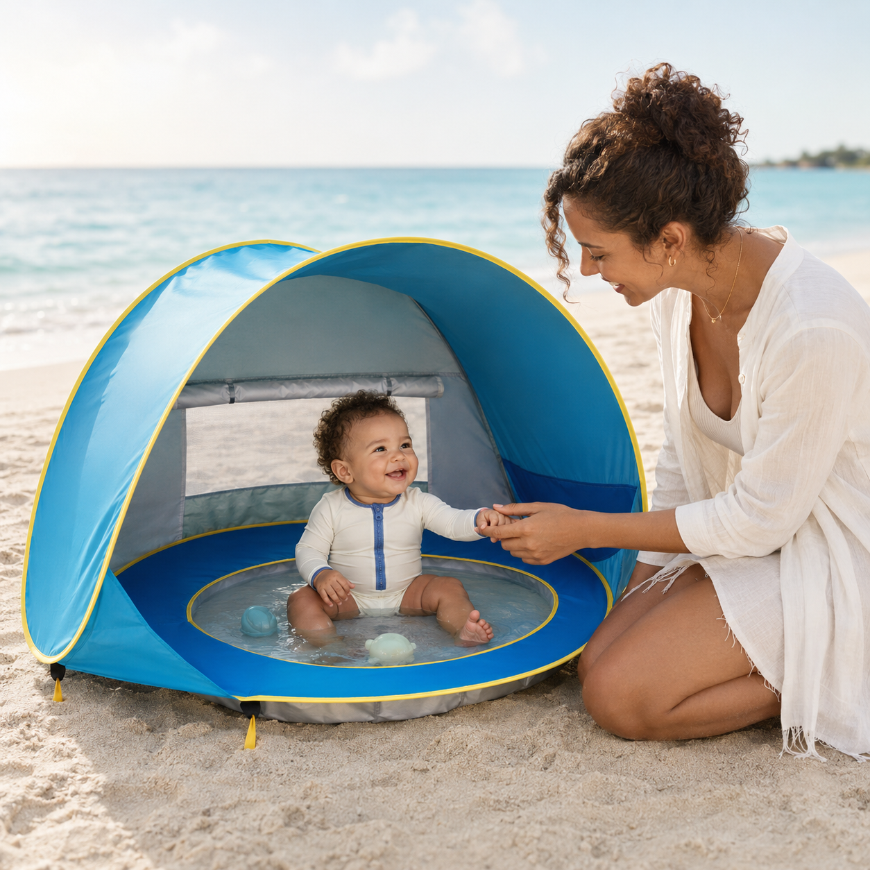 A woman and a baby enjoy a sunny day at the beach, with the woman kneeling beside a blue pop-up beach tent that has a baby pool inside.