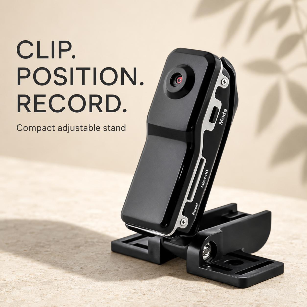 A black compact adjustable camera stand with a small lens detail, shown on a beige textured surface with text reading 'CLIP. POSITION.