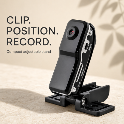 A black compact adjustable camera stand with a small lens detail, shown on a beige textured surface with text reading 'CLIP. POSITION.