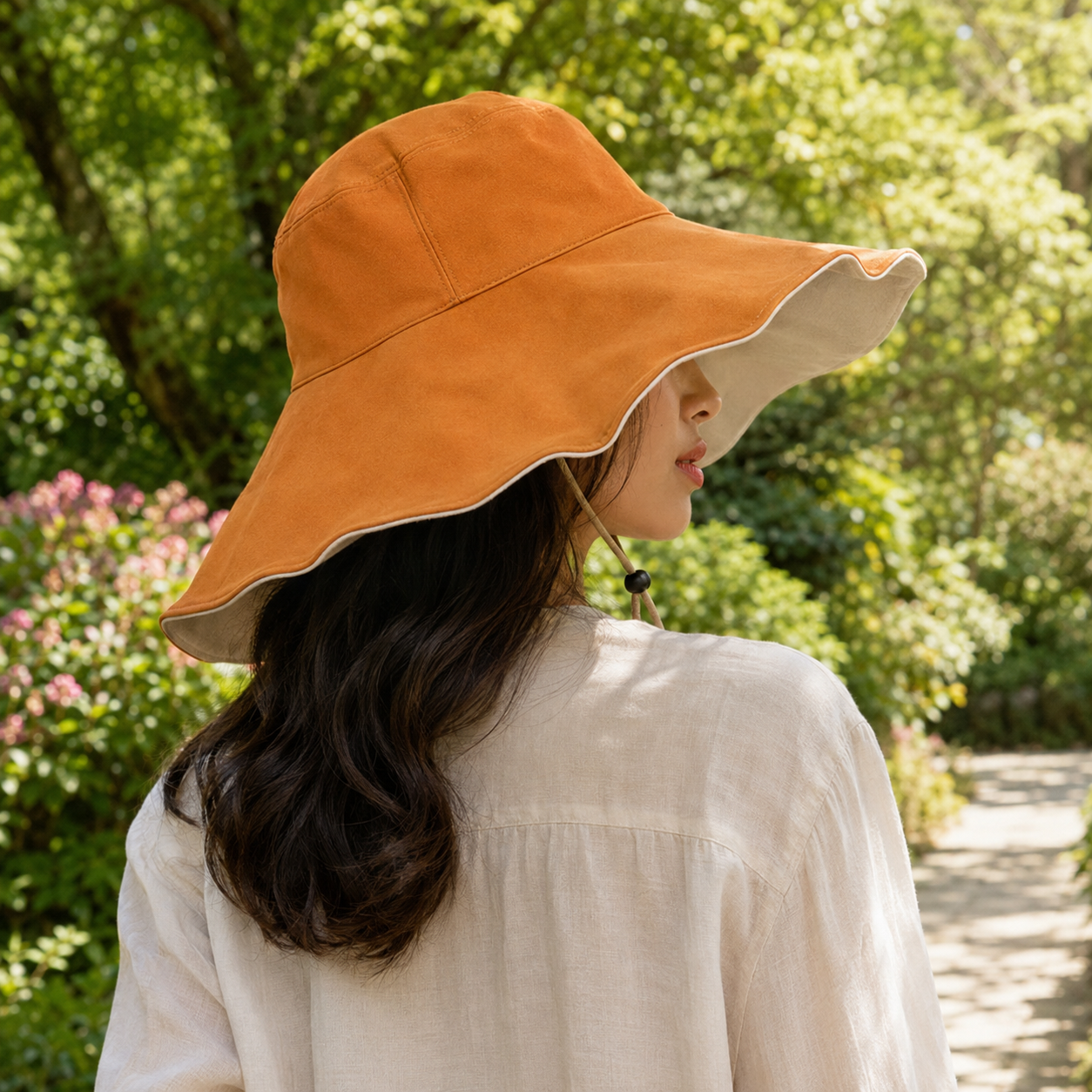 A woman wearing a wide-brimmed orange sun hat with a beige underside and chin strap, photographed outdoors in a lush green garden.