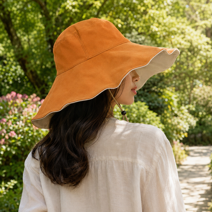 A woman wearing a wide-brimmed orange sun hat with a beige underside and chin strap, photographed outdoors in a lush green garden.